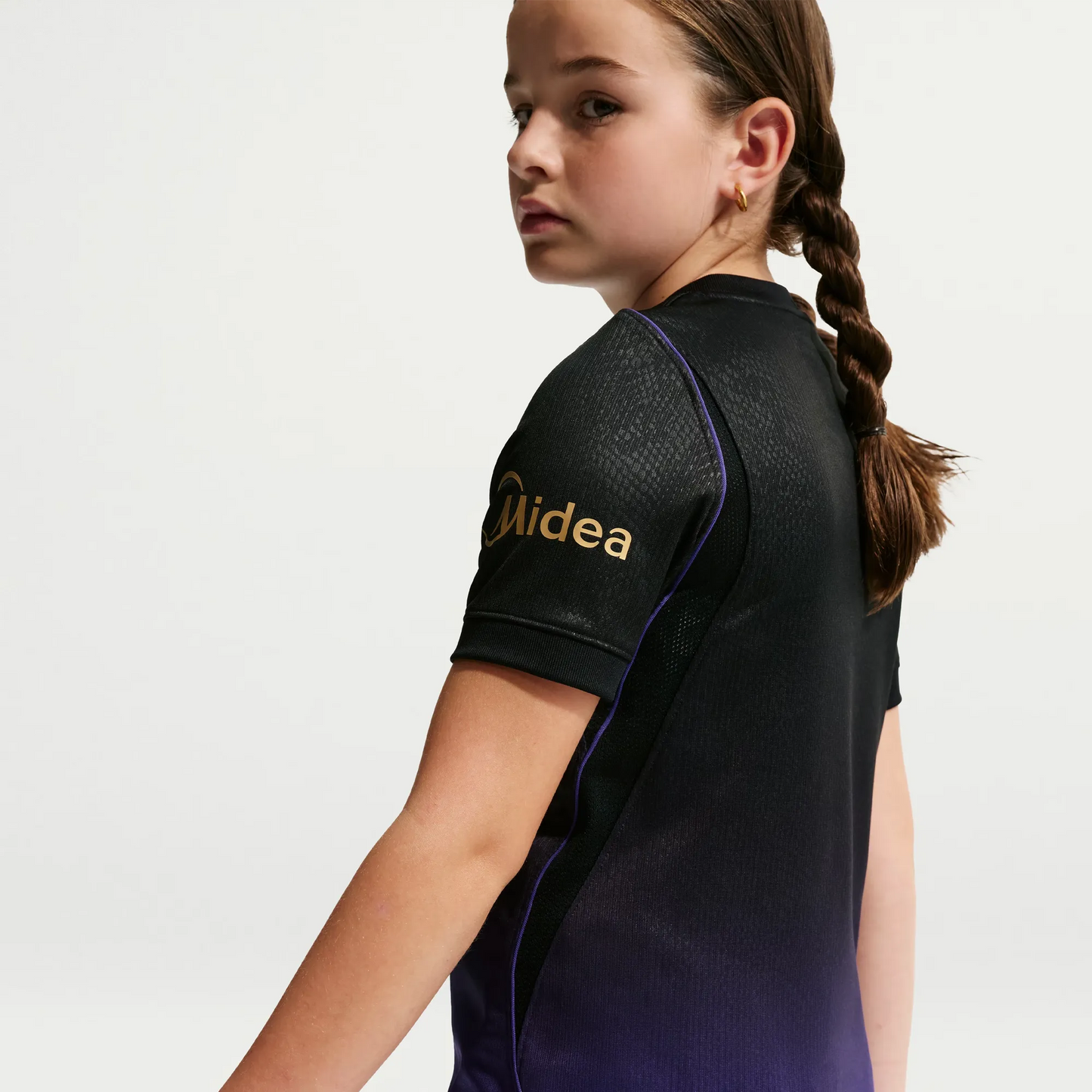 A girl wearing a black short-sleeve athletic jersey with a purple gradient hem and a gold Midea logo on the sleeve, shown from a side-back angle against a light gray studio background.