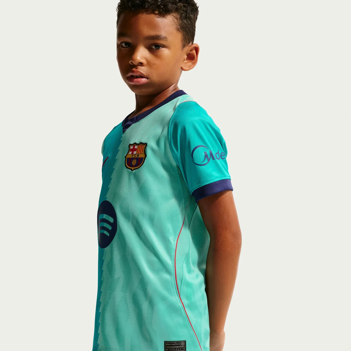A young boy wearing a light turquoise FC Barcelona soccer jersey with dark blue trim, photographed against a clean white studio background.