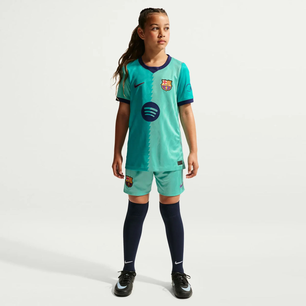 A young girl stands against a light gray studio background wearing a turquoise FC Barcelona soccer kit, including a short-sleeve jersey, matching shorts, navy knee-high socks, and black soccer cleats. Visible branding includes the FC Barcelona crest and Nike logos.