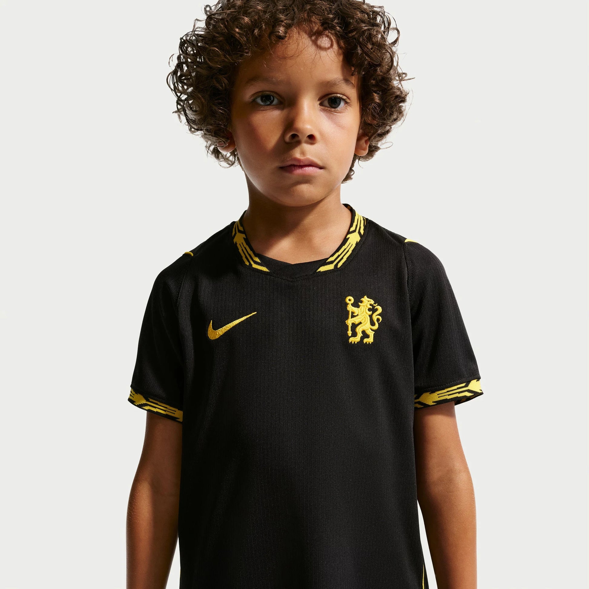 A young boy with curly brown hair wearing a black Chelsea FC home soccer jersey with gold accents, including the Nike swoosh and club crest, against a light gray background.