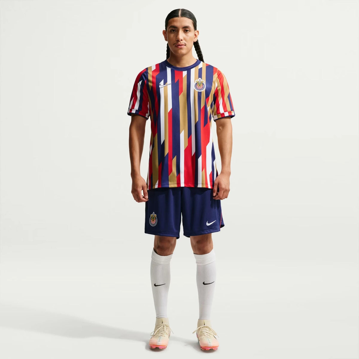 A male soccer player stands against a light gray studio background wearing a Chivas 2026-2027 home white Dri-FIT pre-match top