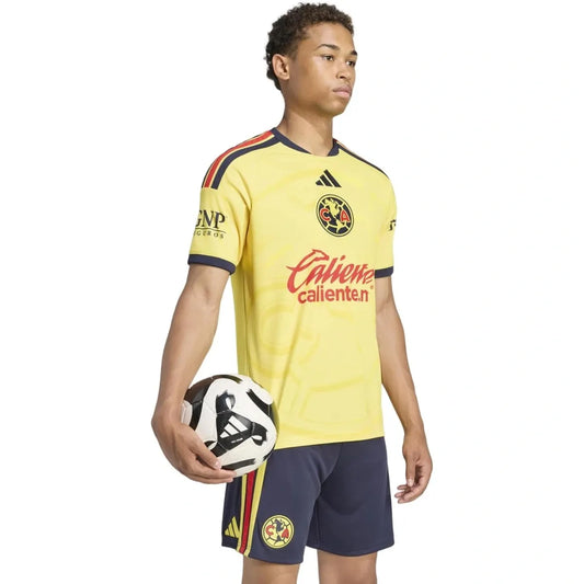 A model wearing a yellow and navy blue Club América soccer jersey and shorts, holding a soccer ball against a white background.