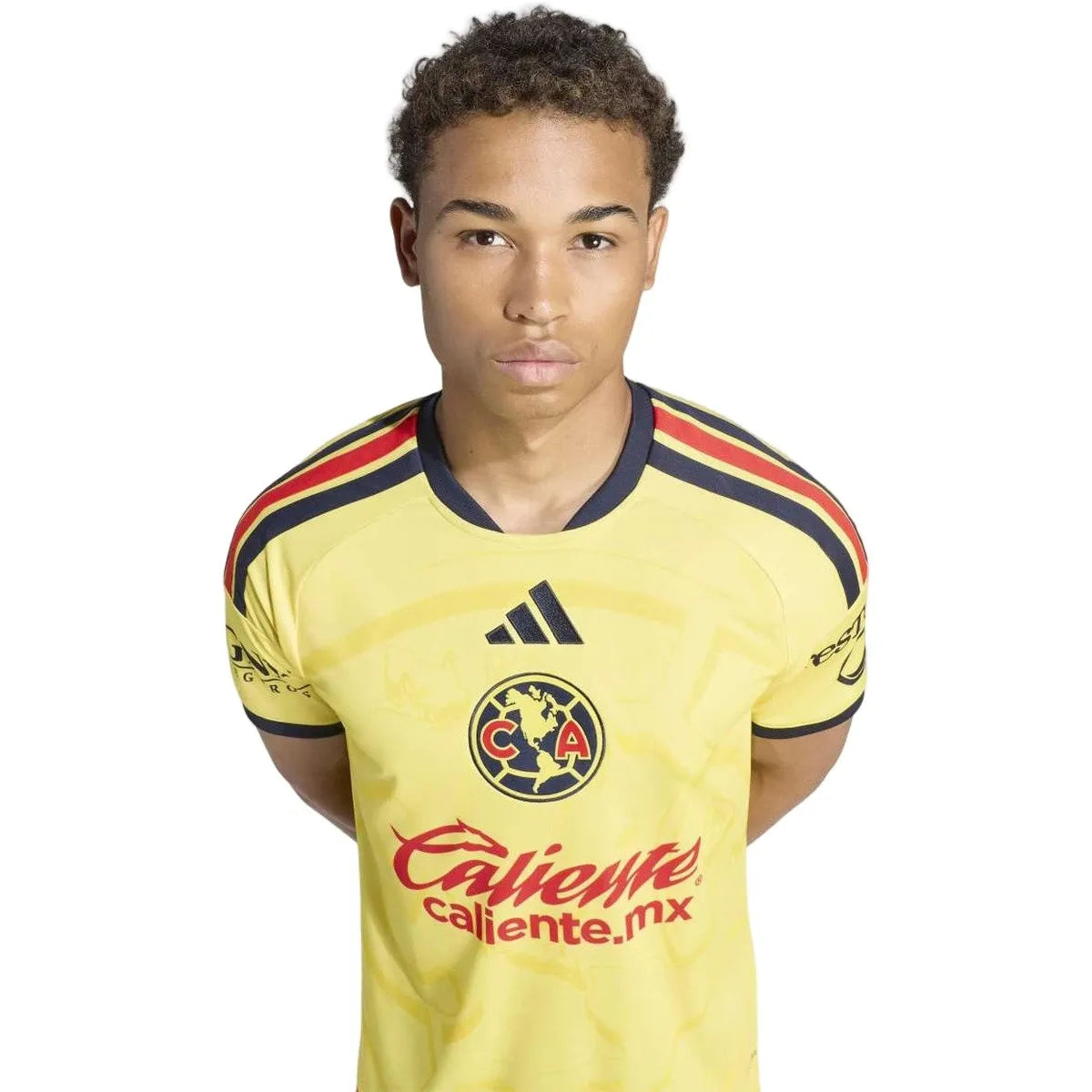 A young man wearing a yellow Club América soccer jersey with navy and red accents, featuring the Adidas logo and Caliente sponsor, against a plain white background.