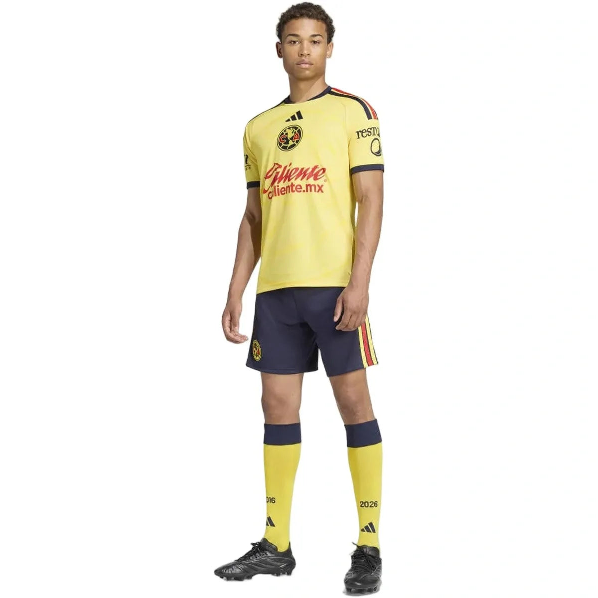 A male model wearing a yellow and navy blue soccer kit, featuring a yellow jersey with red and navy trim, navy shorts with red and yellow stripes, yellow socks, and black cleats, on a solid white background.
