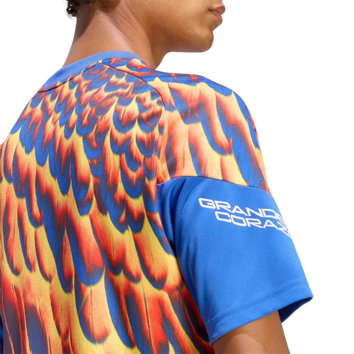 Close-up of a person wearing a short-sleeve t-shirt with a vibrant blue, orange, and yellow feather-like pattern, featuring a solid blue sleeve with white 'GRAND CORAL' text.
