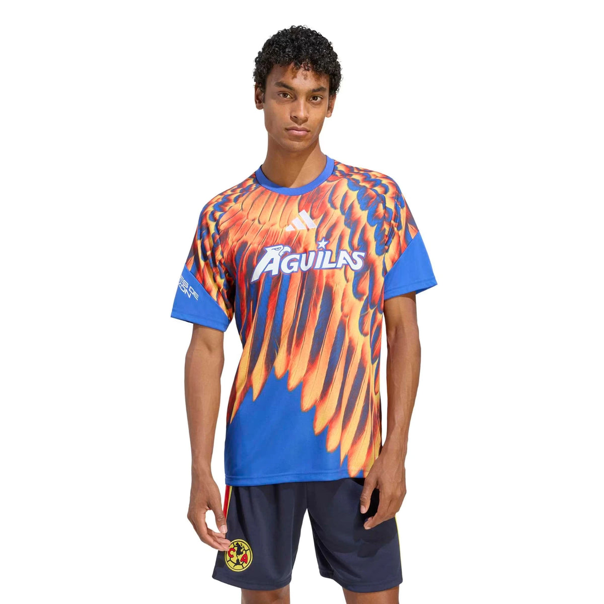 A male model wearing a blue and orange Club América soccer jersey with a wing graphic, navy shorts, and the Adidas logo, set against a plain white background.