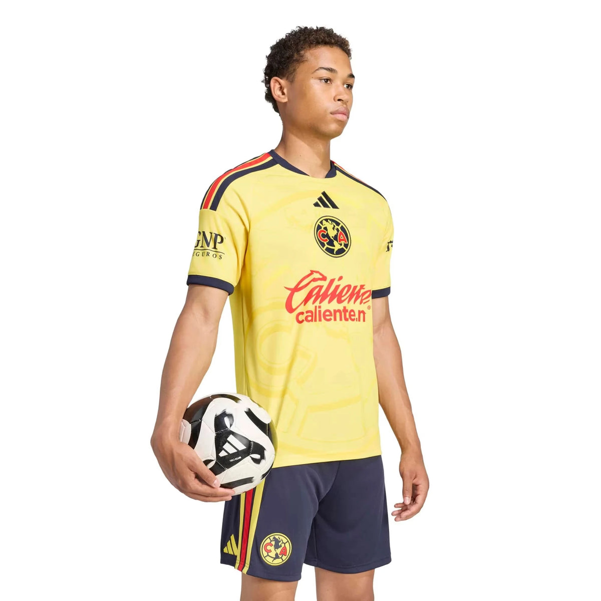 A male model wearing a yellow and navy blue Club América soccer jersey and shorts, holding a soccer ball against a white background.