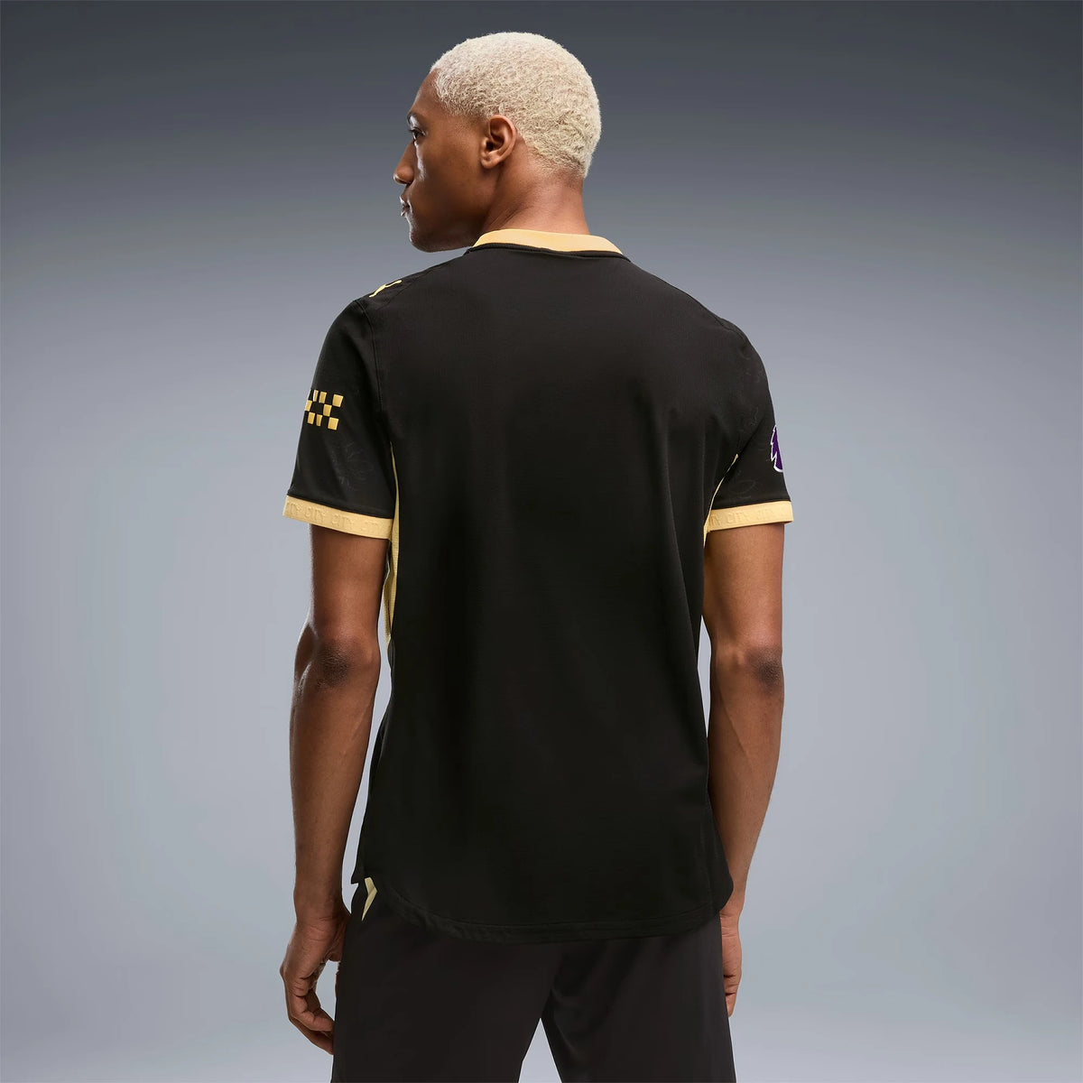 A man with short platinum blonde hair is shown from the back wearing a black short-sleeve t-shirt with gold trim on the collar and cuffs, and a gold checkered pattern on the sleeve. He is standing against a dark gray studio background.