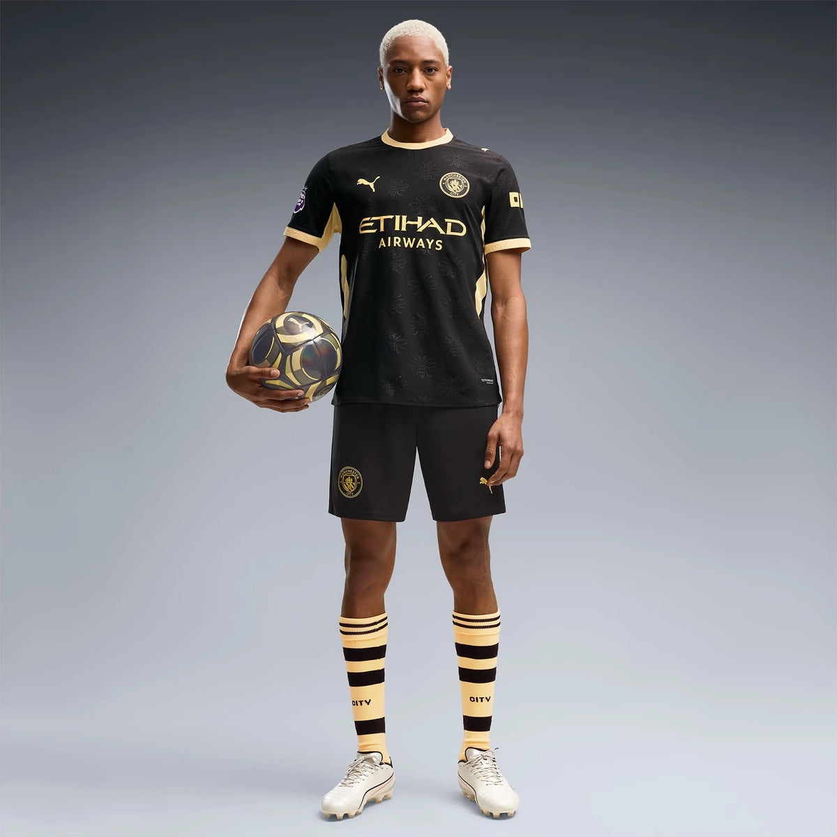 A male model stands against a gray studio background wearing a black Manchester City soccer kit with gold accents, including a short-sleeve jersey, shorts, and striped socks, while holding a soccer ball.