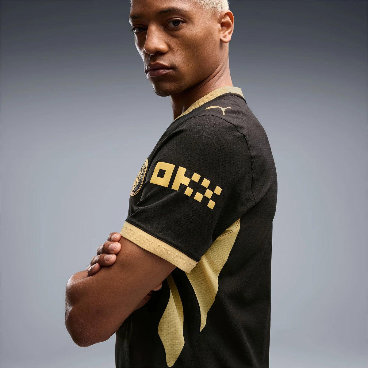 A man wearing a black soccer jersey with gold accents and a Puma logo, photographed against a dark gray studio background.