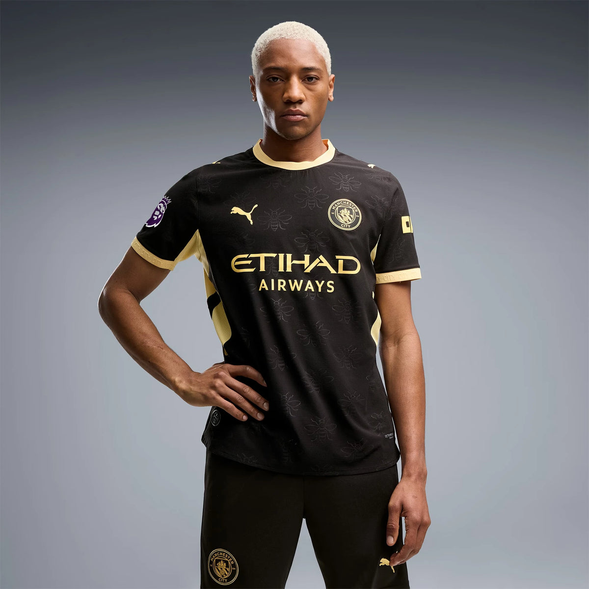A male model wearing a black and gold Manchester City soccer jersey with 'ETIHAD AIRWAYS' and Puma logos, paired with black shorts, against a gray studio background.