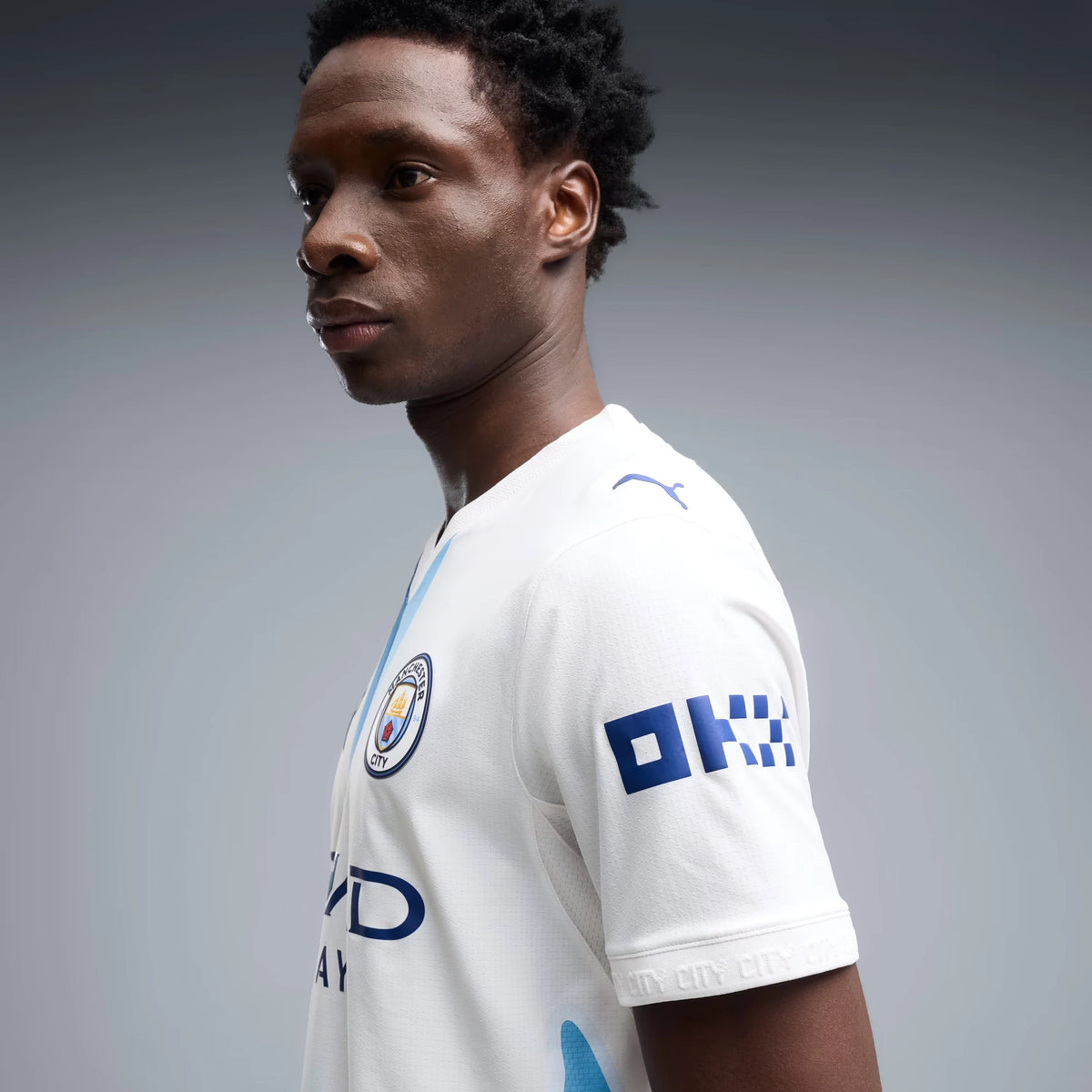 A man in profile wearing a white Manchester City soccer jersey with blue accents, featuring the Puma logo and the OK sponsor logo, set against a solid gray studio background.