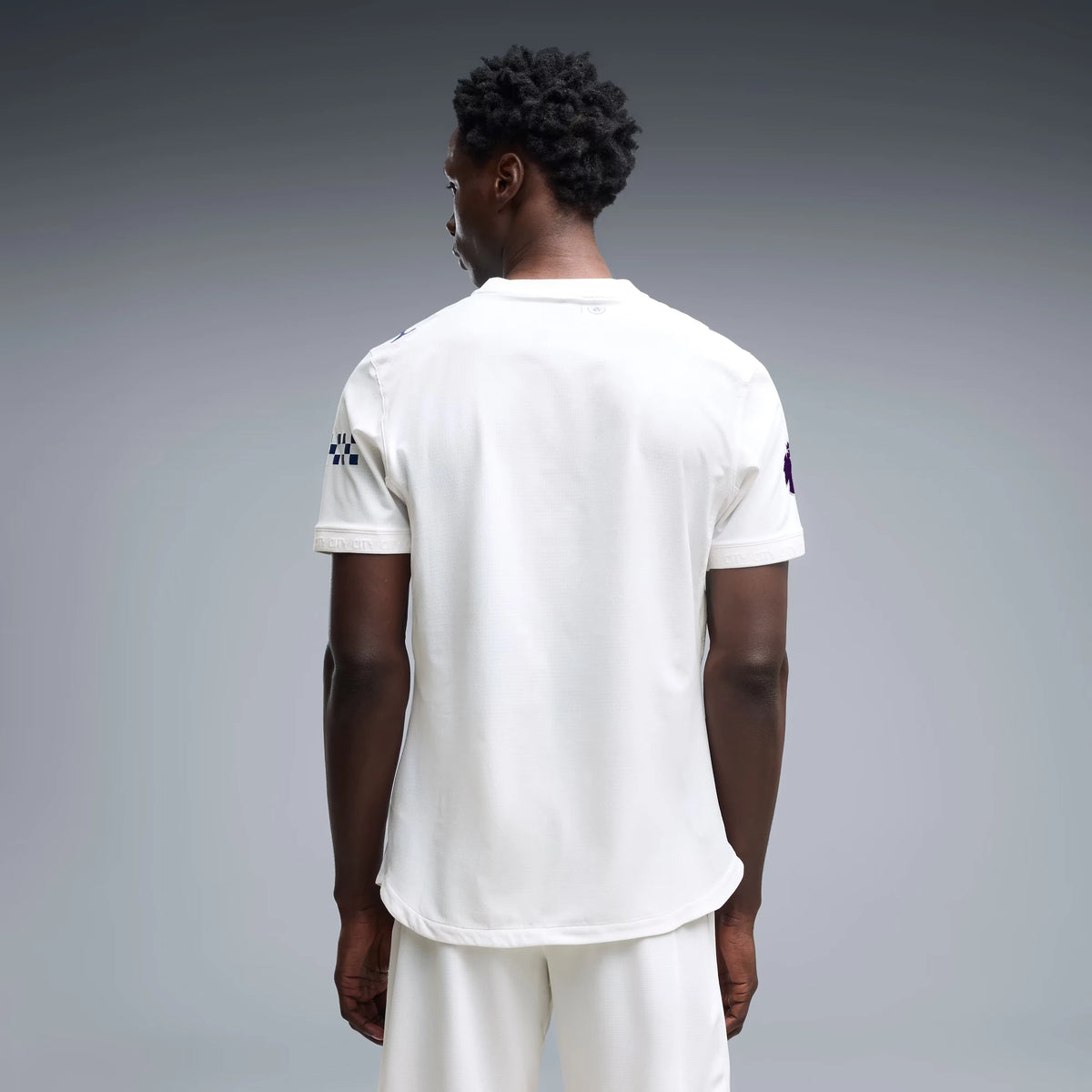 A man viewed from behind wearing a white short-sleeve athletic jersey and matching white shorts against a smooth gray studio background.