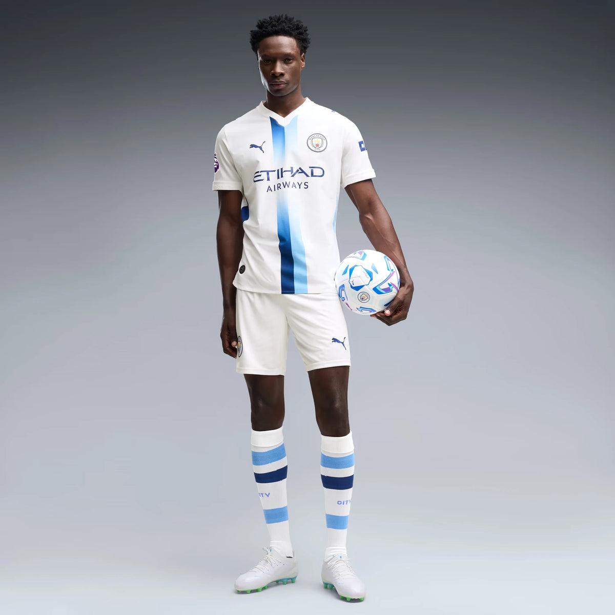 A male soccer player stands against a gray studio background wearing a white Manchester City home jersey with light blue and navy vertical stripes, matching white shorts, white socks with blue stripes, and white soccer cleats. He is holding a blue and white soccer ball.
