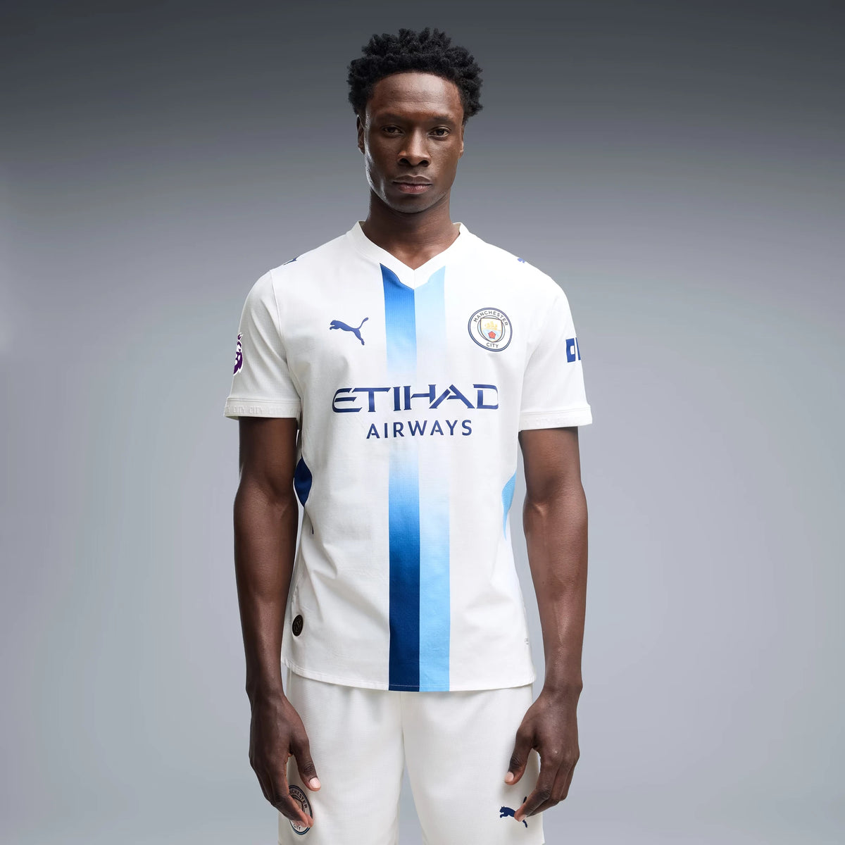 A male model wearing a white Puma Manchester City football jersey with blue vertical stripes, 'ETIHAD AIRWAYS' sponsor text, and the Puma logo, paired with matching white shorts, against a gray studio background.