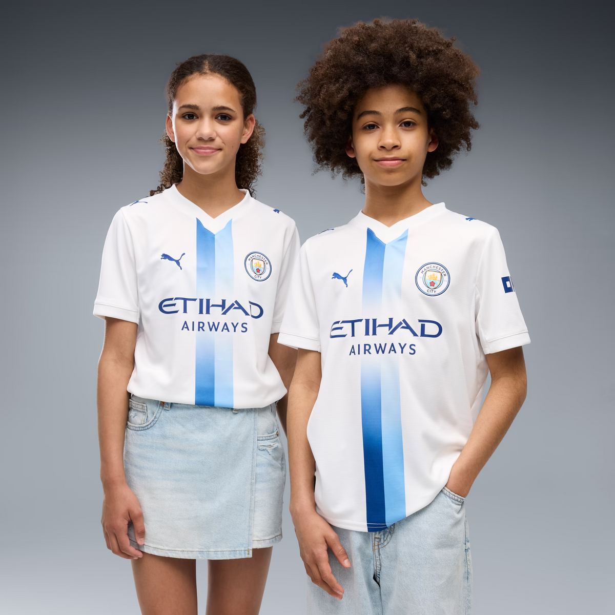 Two children modeling matching white Puma Manchester City football jerseys with light blue vertical stripes and 'ETIHAD AIRWAYS' sponsor text, posed against a gray studio background.