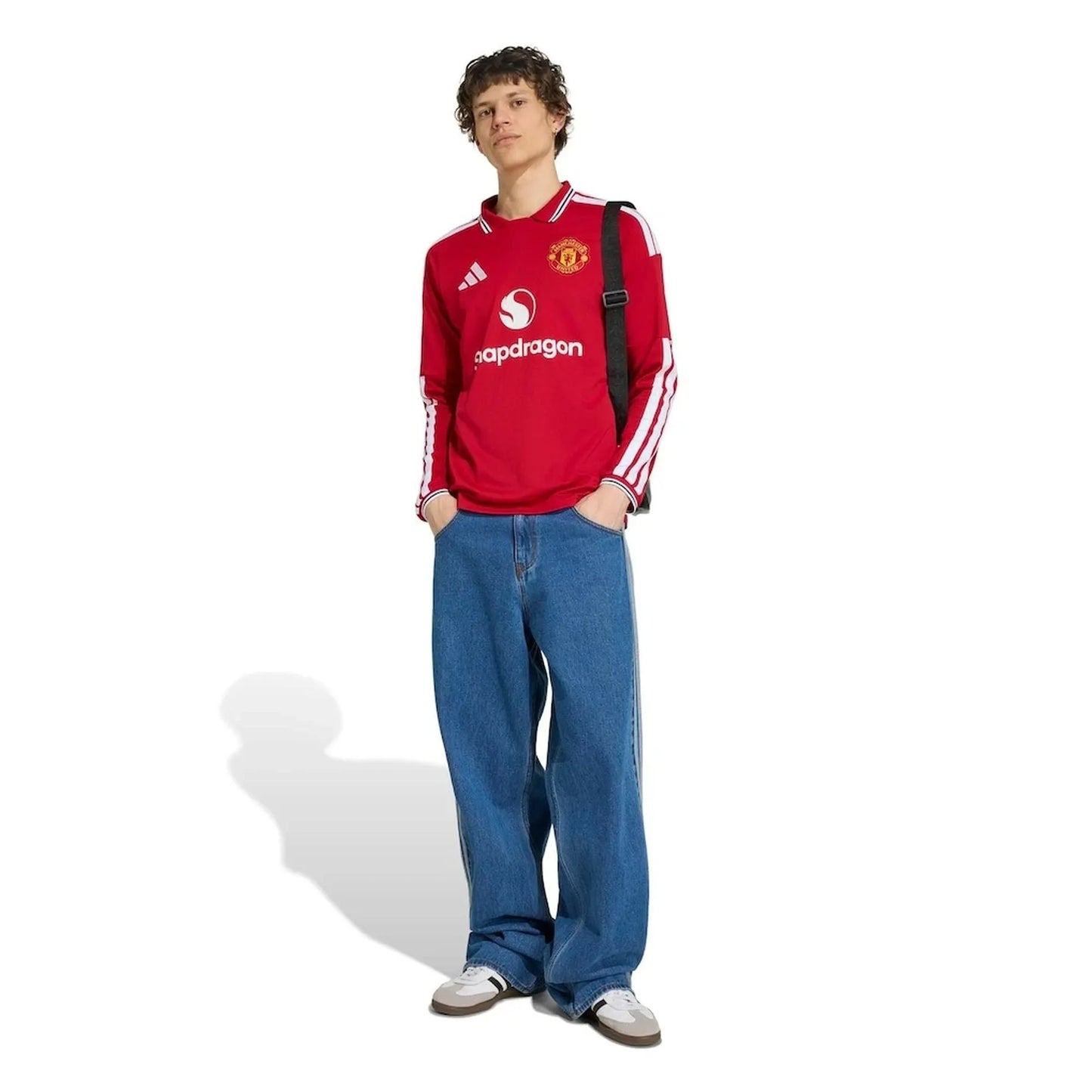 A person modeling a red Manchester United long-sleeve soccer jersey with white Adidas stripes and logos, paired with loose blue jeans and white sneakers, standing against a plain white background.