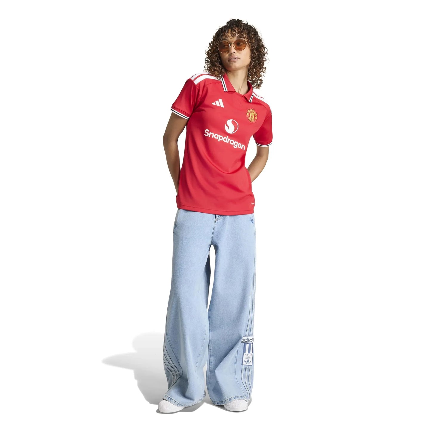 A model wearing a red Manchester United short-sleeve soccer jersey with white trim, featuring the Adidas logo and Snupdragon sponsor, paired with light blue wide-leg jeans and white sneakers, set against a plain white background.