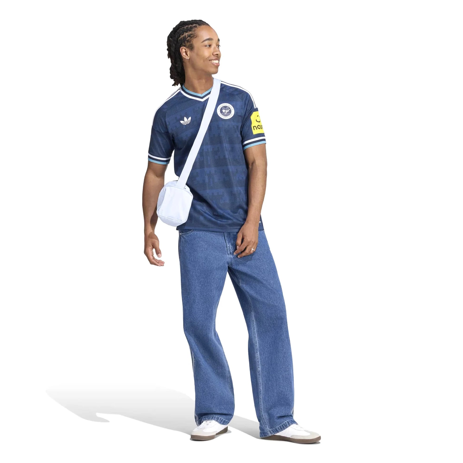 A man modeling a navy blue Adidas soccer jersey with white stripes, paired with wide-leg blue jeans, a white crossbody bag, and white sneakers, set against a plain white background.
