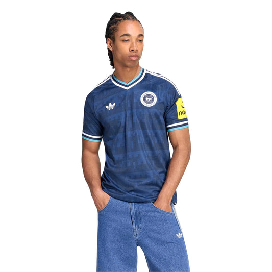 A man wearing a blue patterned Adidas Newcastle United football jersey with white stripes and a yellow sponsor patch on the sleeve, paired with blue jeans, against a plain white background.