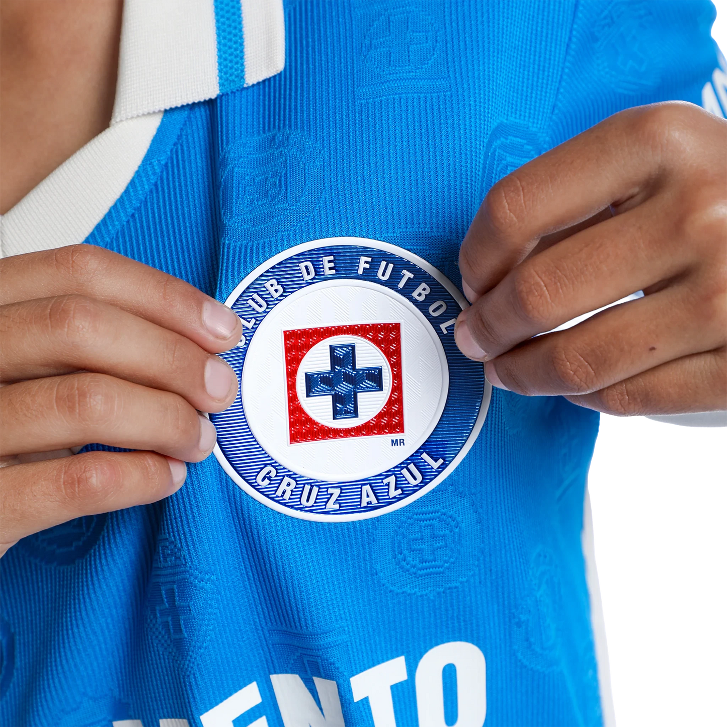A close-up of a person wearing a bright blue Cruz Azul soccer jersey, holding a circular club crest patch in front of the chest.