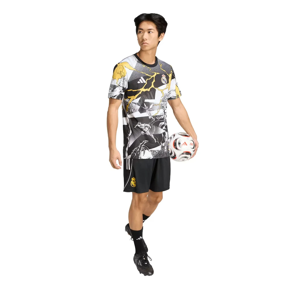 A male model wearing a black, white, and gray soccer jersey with yellow lightning accents, black athletic shorts, black socks, and soccer cleats, holding a soccer ball against a solid white background.
