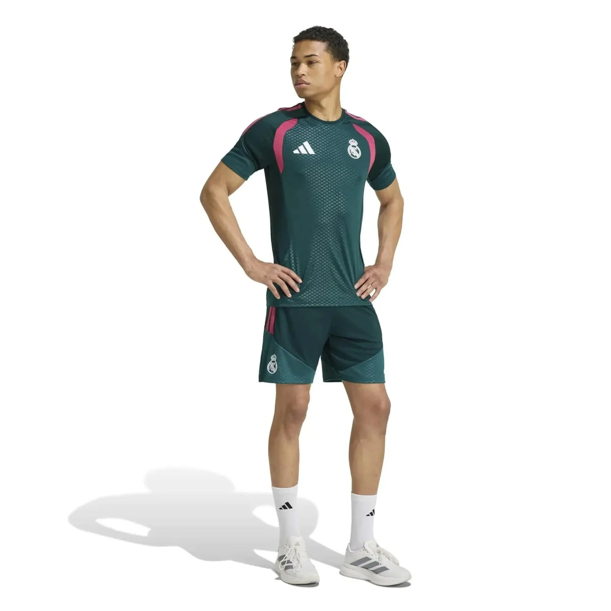 A male model wearing a dark green Real Madrid soccer kit by Adidas, featuring a textured short-sleeve jersey with pink and white accents, matching shorts, white socks, and white Adidas cleats, set against a plain white background.