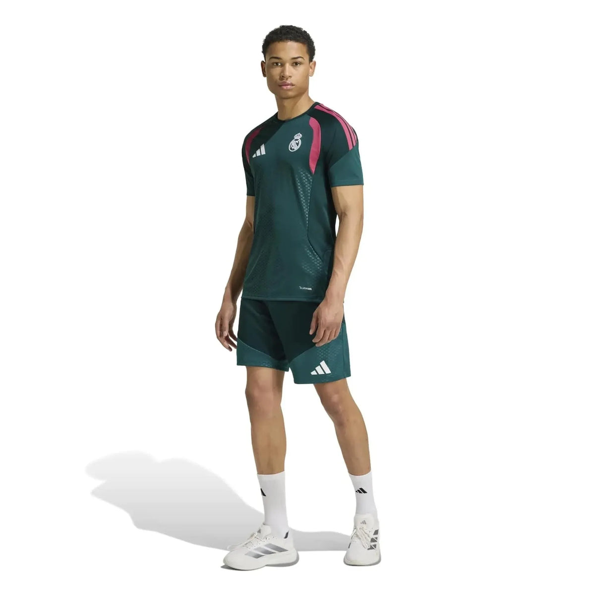 A male model wearing an Adidas Real Madrid training kit, consisting of a dark green short-sleeve jersey with pink and white shoulder stripes and logos, matching dark green shorts, white socks, and white Adidas cleats, set against a plain white background.