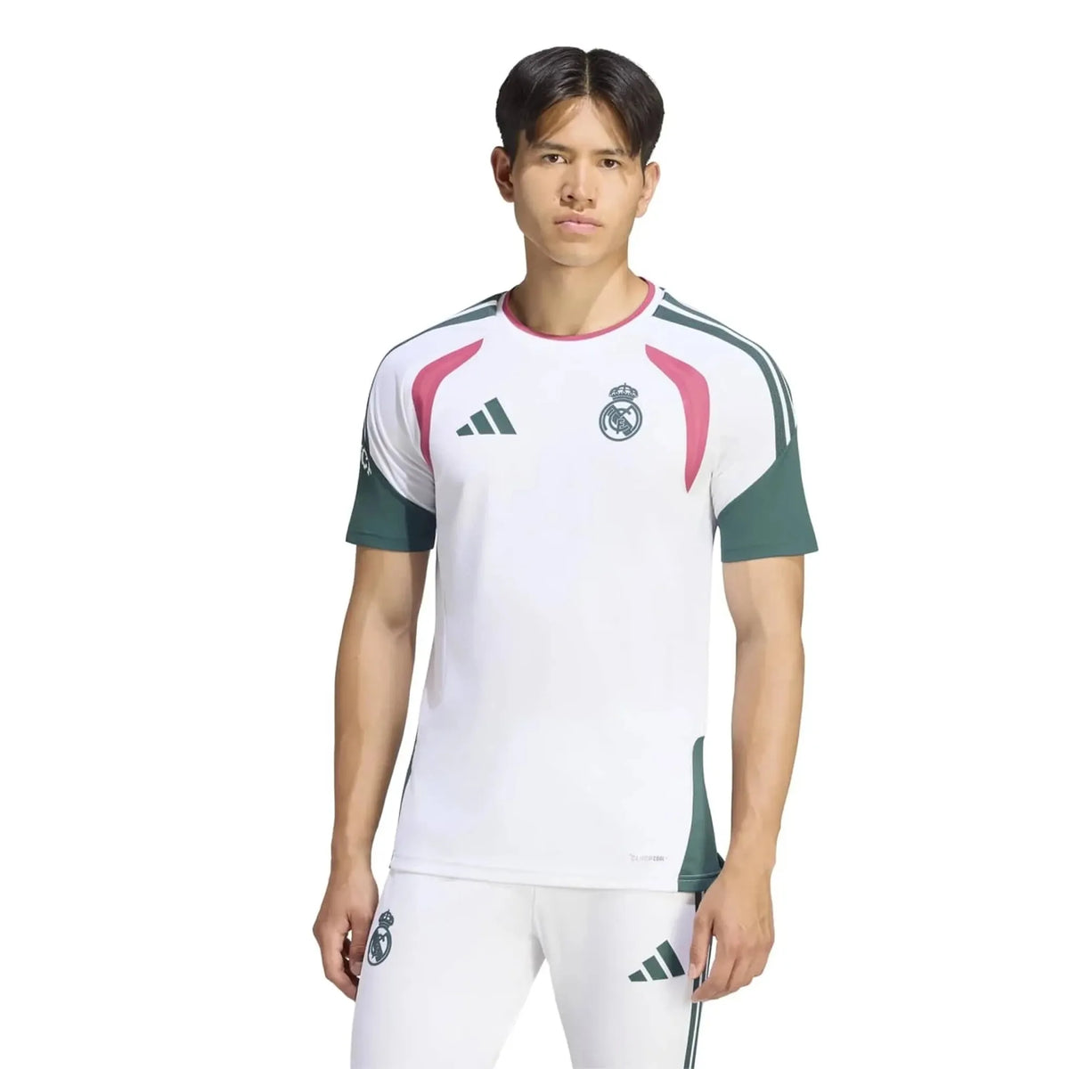 A male model wearing a white Real Madrid soccer jersey with green sleeves and pink accents, paired with matching white shorts, against a plain white background.