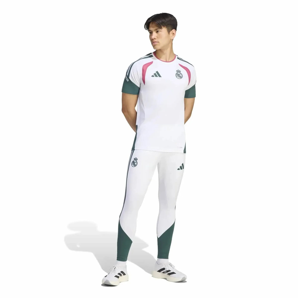 A male model wearing an Adidas Real Madrid athletic tracksuit, featuring a white short-sleeve jersey with green and pink accents, matching white leggings with green panels, and white Adidas cleats, set against a plain white background.