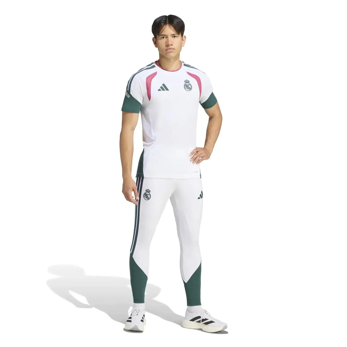 A man modeling a white Adidas Real Madrid soccer kit, featuring a short-sleeve jersey with green and pink accents, matching white and green track pants, and white Adidas cleats, set against a plain white background.