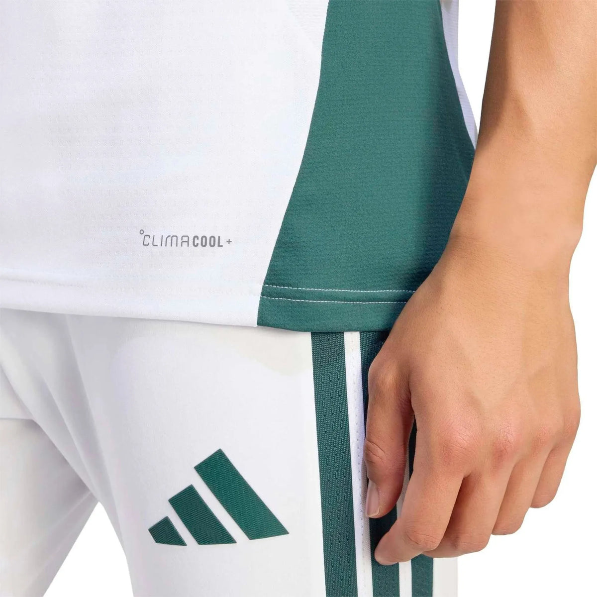 Close-up of a person wearing white Adidas athletic shorts with dark green side stripes and a green Adidas logo, plus a matching white and green top. The photo shows part of the person's hand and torso against a clean white background.
