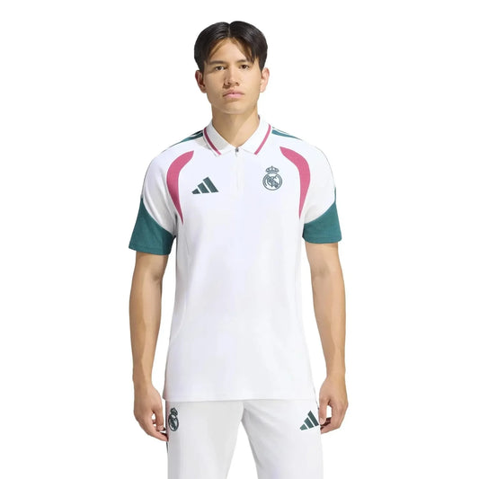 A male model wearing a white Real Madrid polo shirt with green sleeves and pink shoulder accents, paired with white shorts, against a plain white background.