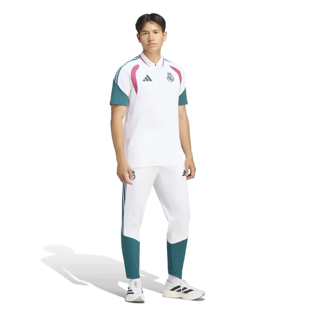 A male model wearing an Adidas Real Madrid soccer kit, featuring a white short-sleeve polo shirt with teal and pink accents, matching white and teal trousers, and white Adidas sneakers, set against a plain white background.