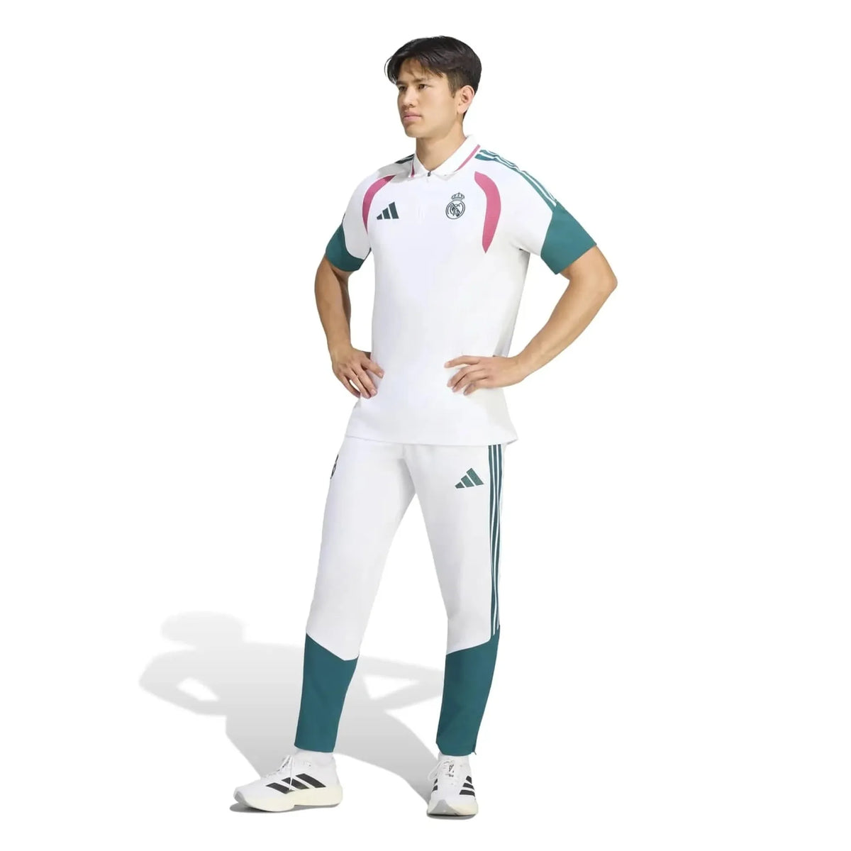 A man modeling a white Real Madrid soccer kit by Adidas, featuring a short-sleeve polo shirt with pink and teal accents, matching white track pants with teal leg panels, and white soccer cleats, set against a plain white background.