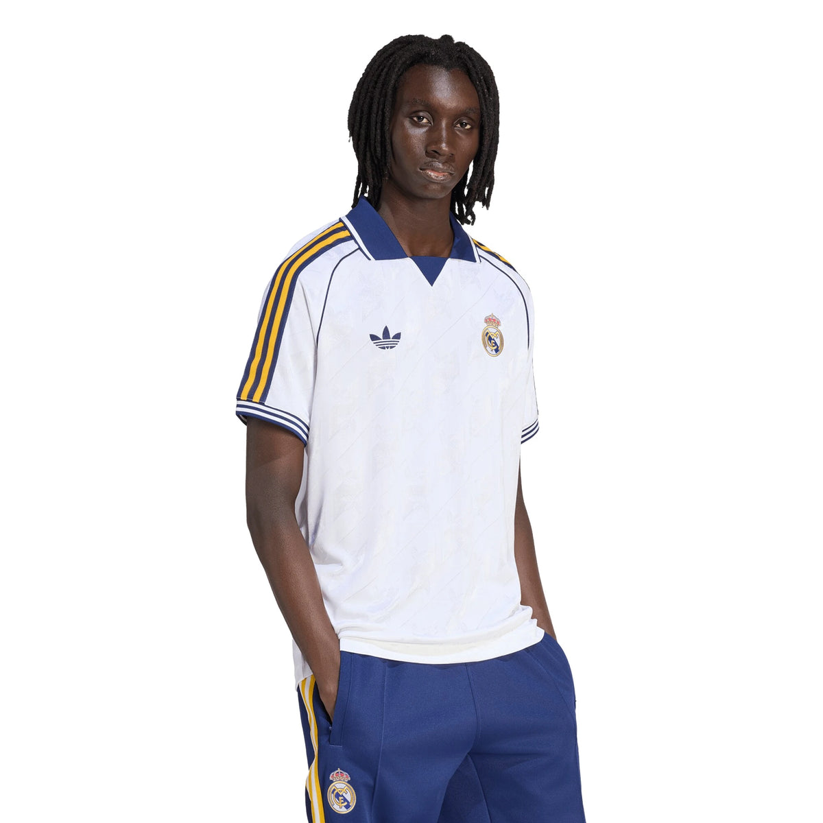 A man modeling a white Real Madrid soccer jersey with blue and yellow stripes on the shoulders and collar, paired with blue shorts, against a plain white background.