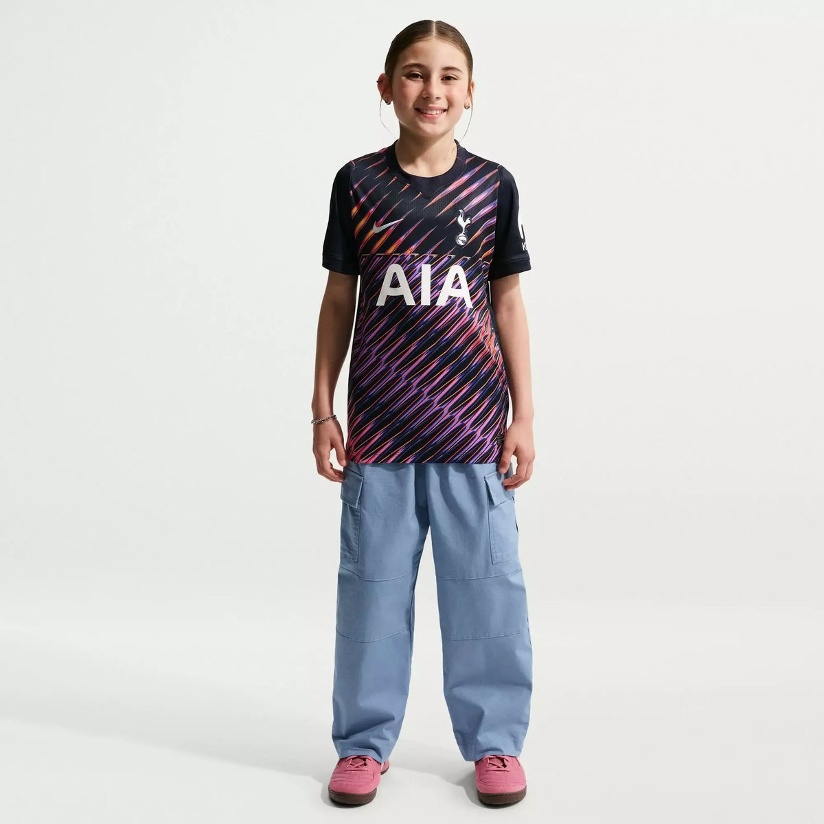 A young girl stands against a light gray studio background wearing a Tottenham Hotspur soccer jersey with a black, purple, and pink diagonal stripe pattern, light blue cargo pants, and pink shoes.