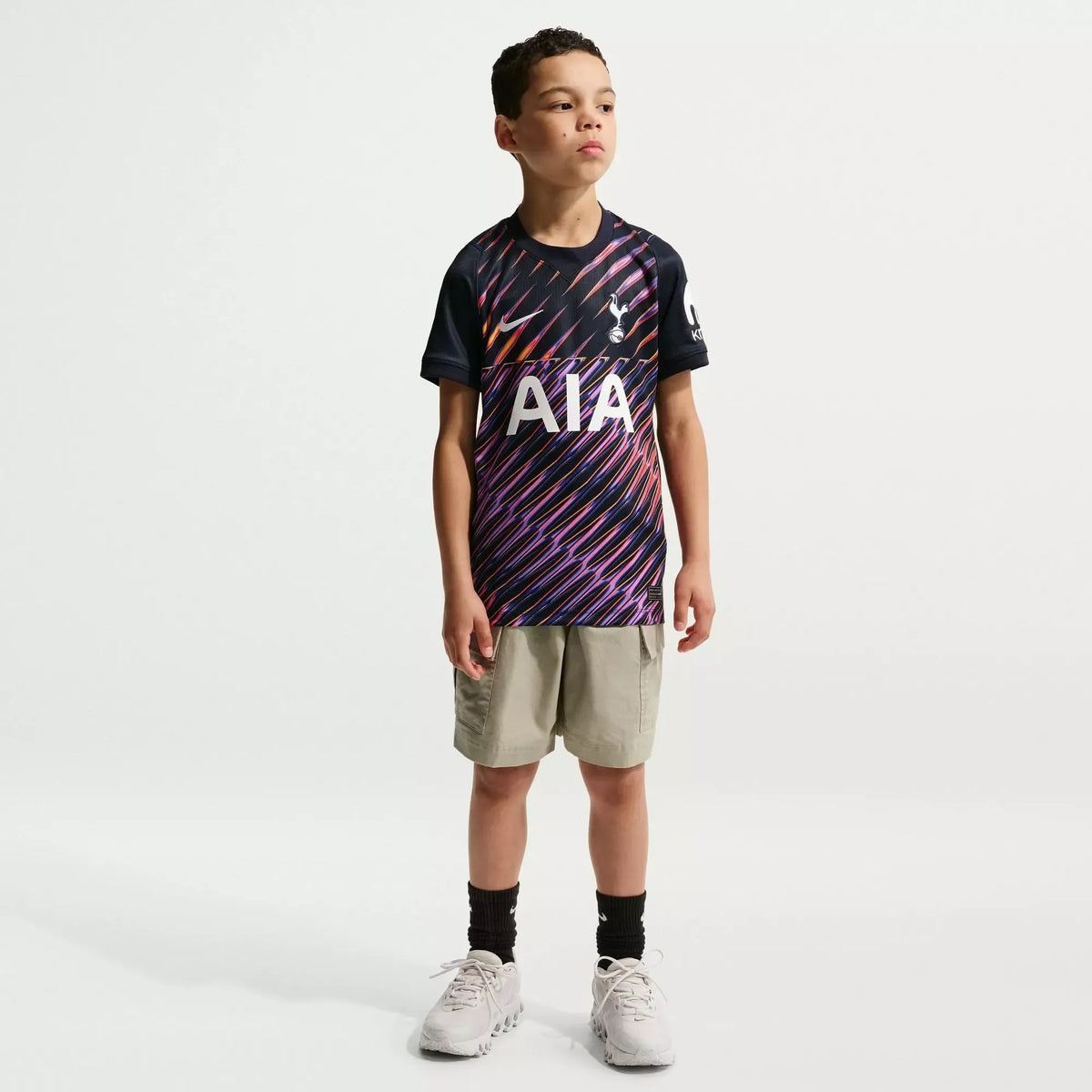 A young boy modeling a Tottenham Hotspur home soccer jersey with a purple and orange diagonal stripe pattern, paired with khaki shorts, black socks, and white sneakers against a light gray studio background.