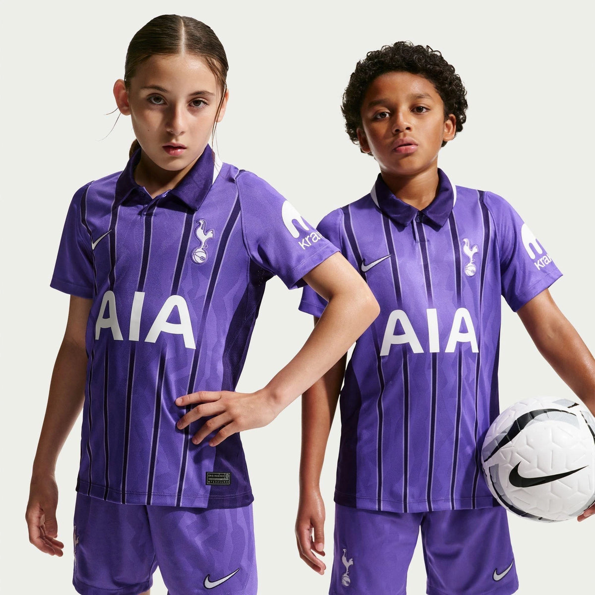 Two children modeling a purple Tottenham Hotspur football kit with vertical stripes, featuring the AIA sponsor logo on the front, Nike logos, and a white soccer ball on a light gray background.