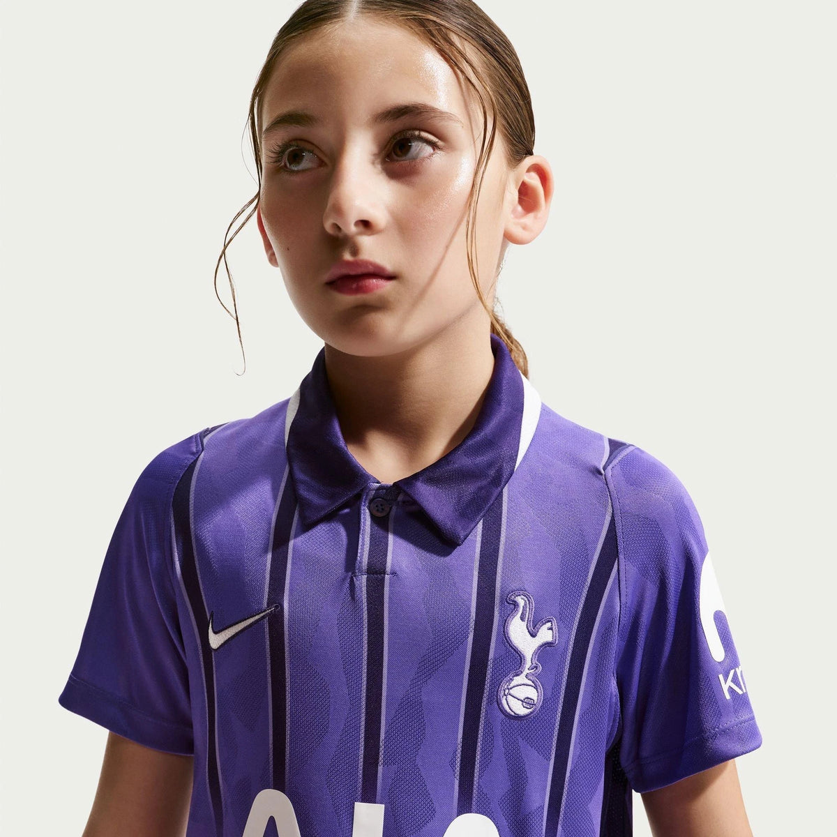 A young girl wearing a purple and navy striped Tottenham Hotspur soccer jersey with a white Nike logo and club crest, photographed against a light gray background.