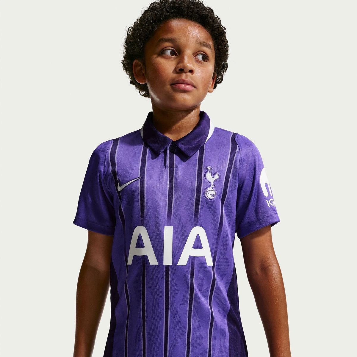 A young boy wearing a purple and black striped Tottenham Hotspur soccer jersey with the AIA sponsor logo, Nike swoosh, and cockerel crest, set against a solid light gray background.