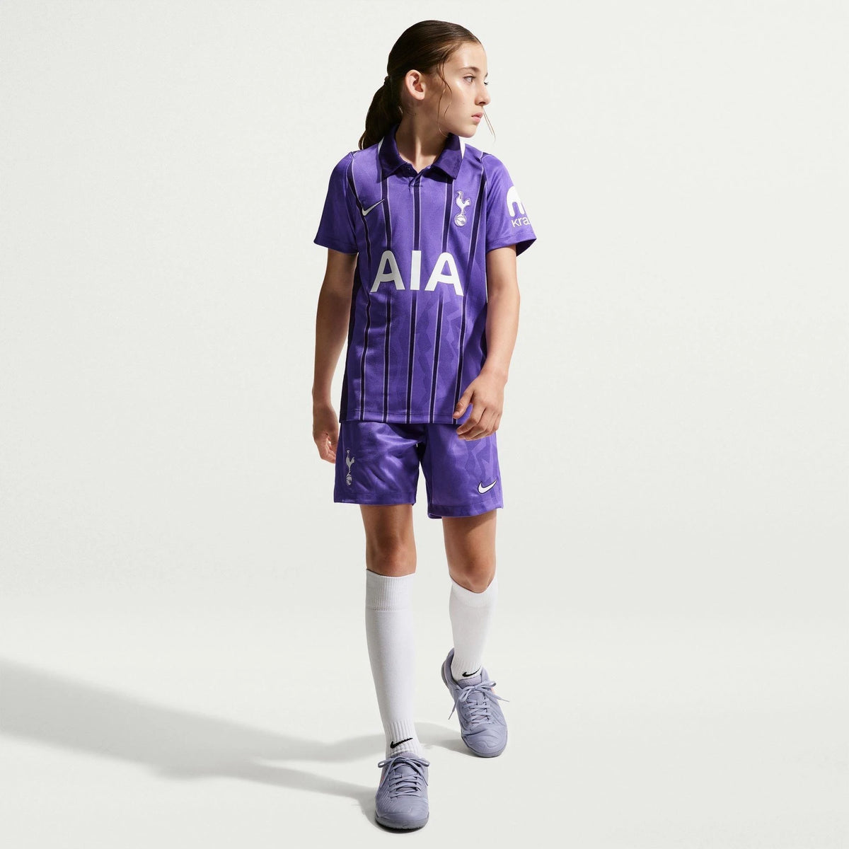 A young girl modeling a purple Tottenham Hotspur football kit, featuring a short-sleeve polo shirt and matching shorts with white Nike logos, white socks, and grey Nike cleats, set against a plain light grey studio background.